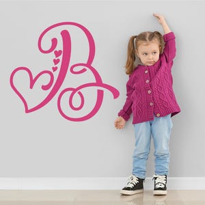 May include: A pink letter "B" with a heart design on a grey wall. The letter is next to a young girl wearing a pink cardigan and blue jeans, measuring her height.