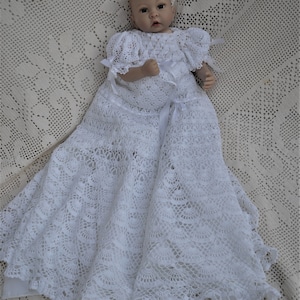 May include: A white, handmade crochet baby christening gown. The gown features a detailed openwork pattern, short sleeves, and delicate ribbon accents. The long skirt flares out, creating a flowing silhouette. The baby doll is lying on a cream-colored crocheted blanket.