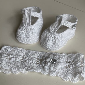 Puede incluir: Zapatos de bebé de crochet blanco con una correa en T y una diadema de crochet blanco con un adorno de flor y un strass.