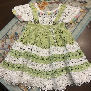 May include: A handmade crochet dress in white and light green. The dress features a detailed openwork design, with a white bodice and a skirt with alternating green and white stripes. The dress has small butterfly accents and a delicate, scalloped hemline.