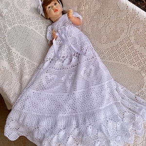 Könnte beinhalten: Weißes gehäkeltes Babykleid mit einem Satinband und einem passenden gehäkelten Stirnband. Das Kleid hat einen gewellten Saum und ein zartes Spitzenmuster.