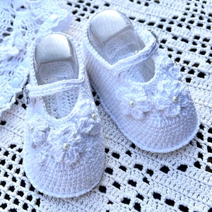 May include: White crocheted baby shoes with a flower design and pearl accents.