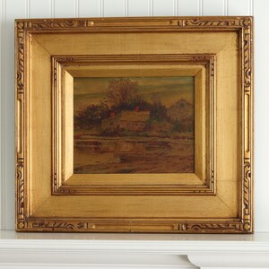 May include: A framed landscape painting with a gold-colored ornate frame. The painting depicts a rural scene with a house, trees, and a body of water. The artwork is displayed on a white mantelpiece against a white wall.