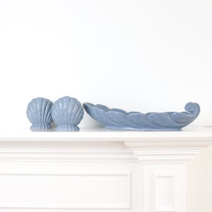 Può includere: Un piatto in ceramica blu chiaro a forma di conchiglia e due portacandele abbinati a forma di conchiglia. Il piatto ha un design a forma di foglia e un manico arricciato. Gli oggetti sono esposti su una superficie bianca.