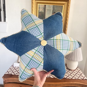 May include: A star-shaped pillow with a patchwork design. The pillow features denim blue fabric and plaid patterns in shades of blue, yellow, and green. A yellow button is in the center. The edges are trimmed with a yellow piping.
