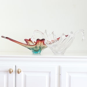May include: Two decorative glass bowls. One is clear, with a wavy, open design. The other bowl is a vibrant mix of colors, transitioning from red to teal. Both bowls are displayed on a white surface.