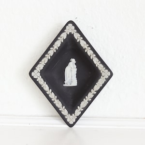 May include: Black diamond-shaped dish with a white floral and leaf border. A white relief figure of two people is in the center. The dish is likely ceramic or porcelain.