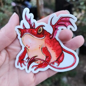 May include: A red and orange cartoon frog with a horn, wings, and a tail. The frog is sitting on its hind legs and has a friendly expression.