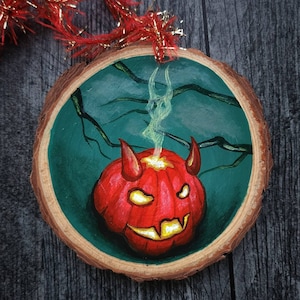 May include: A Halloween-themed wooden ornament featuring a red pumpkin with devil horns and a glowing face. The pumpkin is set against a teal background with dark branches. The ornament has a natural wood edge and a red and gold hanging ribbon.