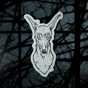 May include: Black and white line art illustration of a dog's head with large, staring eyes. The dog has a long snout and pointy ears. The illustration is on a white background.