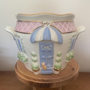 May include: A white ceramic house-shaped planter with a pink and blue roof, blue door, and a small cat figurine. The planter has gold trim and is on a wooden stand.