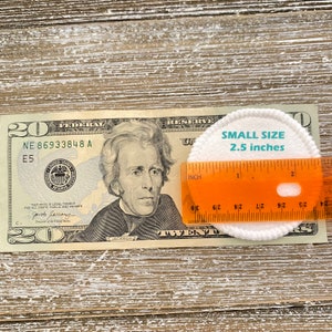 May include: A white cotton round pad measuring 2.5 inches in diameter, placed on top of a twenty dollar bill with a ruler beside it. The ruler is orange and has markings in inches.