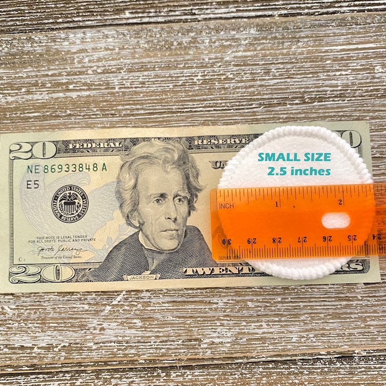 May include: A white cotton round pad measuring 2.5 inches in diameter, placed on top of a twenty dollar bill with a ruler beside it. The ruler is orange and has markings in inches.