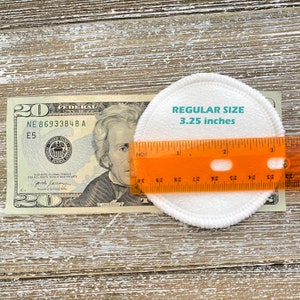May include: A white cotton round makeup remover pad measuring 3.25 inches in diameter, next to a twenty dollar bill and a ruler.
