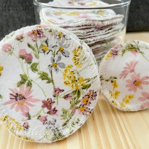以下が含まれることがあります： 白い背景にピンクと黄色の花と緑の葉が描かれた、円形の再利用可能な布製フェイシャルパッドのスタック。各円形パッドの縁は、耐久性を高めるために白いオーバーロックステッチで仕上げられています。
