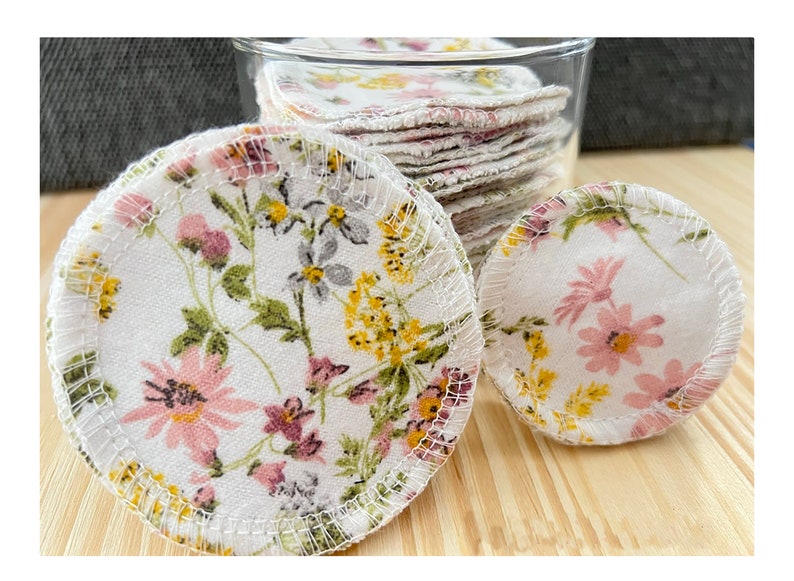 May include: A close-up of reusable cotton rounds with a floral print. The rounds are white with pink and yellow flowers. The rounds are in a glass jar and on a wooden surface.