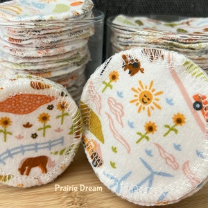 May include: Two glass jars filled with reusable cotton rounds. The rounds are white with different colorful patterns, including a sun, sunflowers, and a farm scene. The text "Prairie Dream" is visible on the wooden surface.