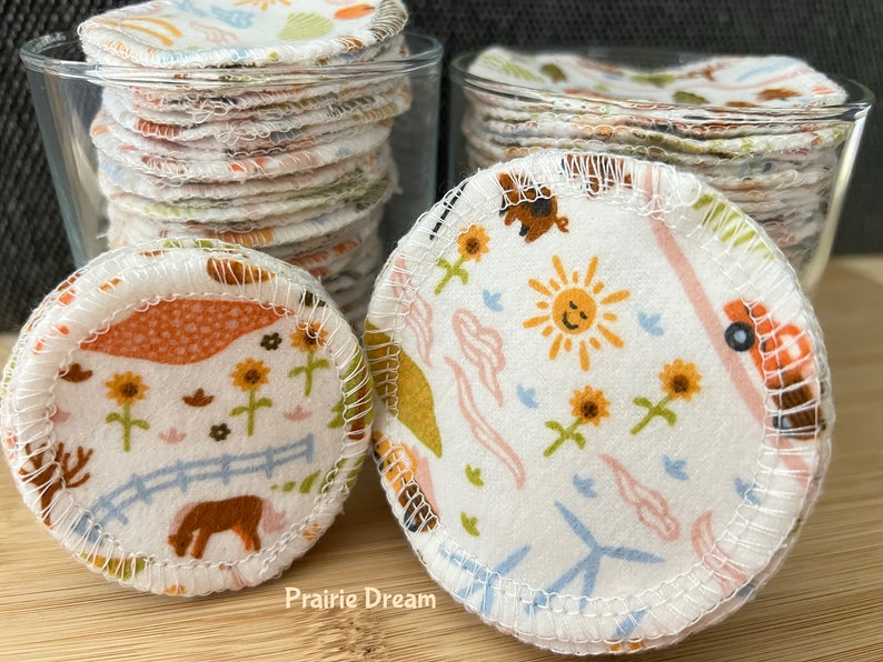 May include: Two glass jars filled with reusable cotton rounds. The rounds are white with different colorful patterns, including a sun, sunflowers, and a farm scene. The text "Prairie Dream" is visible on the wooden surface.