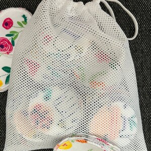 May include: White mesh laundry bag with a drawstring closure. The bag is filled with round, white cotton makeup remover pads with floral patterns.