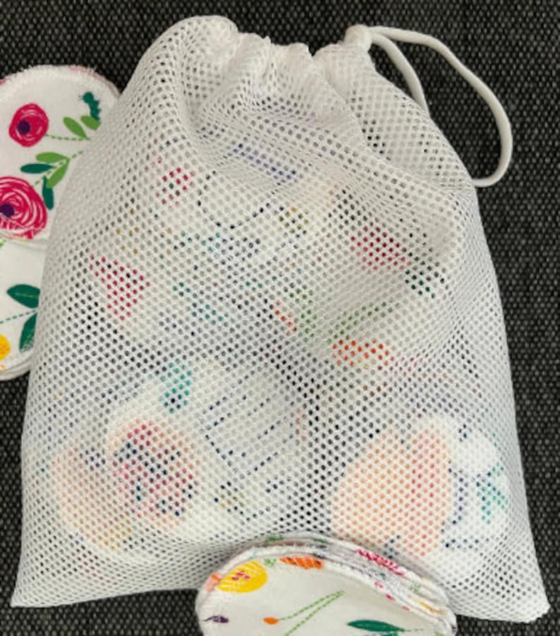 May include: White mesh laundry bag with a drawstring closure. The bag is filled with round, white cotton makeup remover pads with floral patterns.