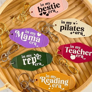 May include: A collection of retro-style keychains in various colors, each with a unique phrase in white. Phrases include "in my bestie era," "in my pilates era," and "in my reading era." The keychains are displayed on a woven tray.