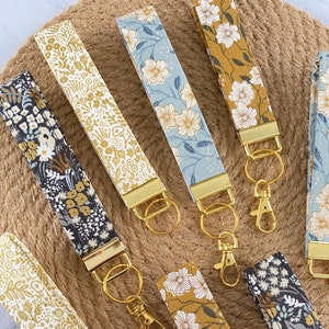May include: A collection of floral patterned key fobs in various colors, including gold, blue, and mustard yellow. Each fob features a gold-colored metal clasp and key ring. The fabric designs showcase white flowers and foliage.