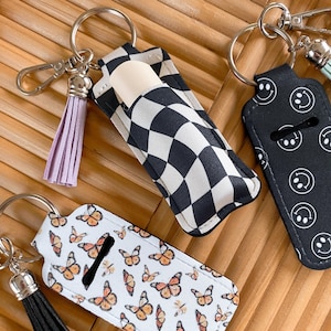 May include: Three lip balm holders with keychains. One has a black and white checkered pattern, another features a butterfly design, and the third has a smiley face pattern. Each holder has a tassel in a different color.