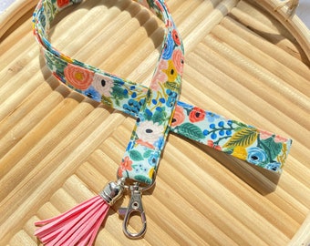 May include: A fabric lanyard featuring a light blue background with a colorful floral pattern of orange, pink, yellow, and dark blue flowers and green leaves. The lanyard includes a silver metal lobster clasp and a bright pink faux leather tassel.