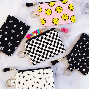 May include: A collection of small, zippered coin purses in various patterns. Designs include a pink background with yellow smiley faces, a black and white checkerboard, a black background with white stars, and a white background with black plus signs.