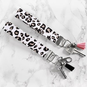 May include: Two key wristlets with a white background and a black and tan leopard print pattern. Each wristlet has a silver-colored metal clasp and key ring. One wristlet has a pink tassel, and the other has a black tassel.