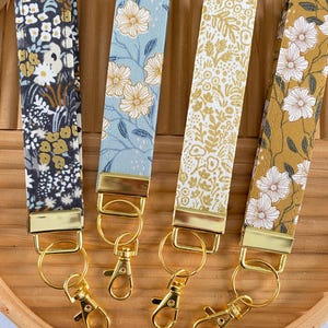 May include: Four fabric wrist keychains with floral patterns in navy, blue, white, and gold. Each keychain has a gold-colored metal clasp and ring. The keychains are arranged on a wooden tray.