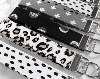 Può includere: Una collezione di portachiavi in tessuto con vari motivi, tra cui stampa leopardata, pois e croci. I portachiavi sono in tonalità di nero, bianco e grigio, con anelli e finiture argentate.