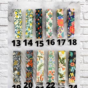 May include: A selection of fabric key fobs with various floral and botanical patterns. Each fob is numbered, with the text "CHOOSE YOUR FABRIC" at the top. The key fobs are displayed against a white and gray background.