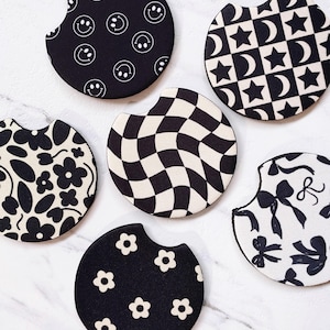 May include: A collection of six car coasters with various black and white patterns. Designs include smiley faces, stars and moons, a checkered pattern, floral patterns, and bows. These coasters are designed to fit in a car's cup holder.