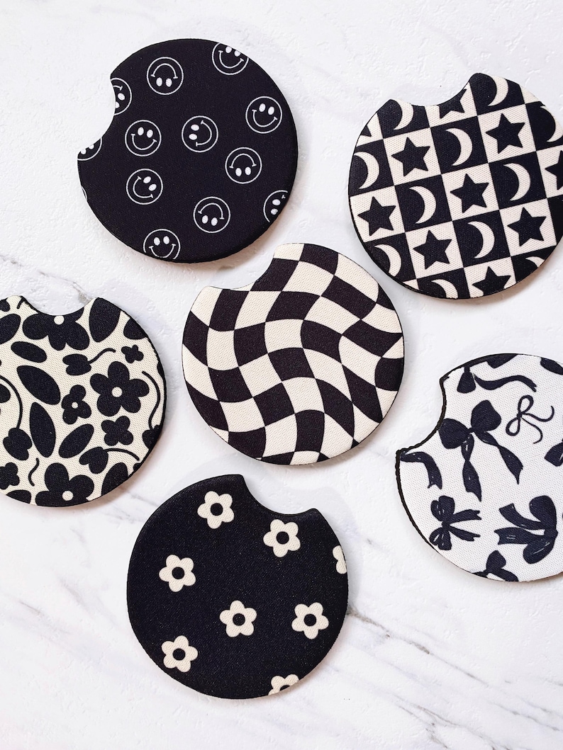 May include: A collection of six car coasters with various black and white patterns. Designs include smiley faces, stars and moons, a checkered pattern, floral patterns, and bows. These coasters are designed to fit in a car's cup holder.
