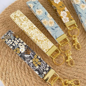 May include: Several fabric key fobs with floral patterns in various colors, including mustard yellow, light blue, and black. Each fob has a gold-colored metal clasp and ring for attaching keys. The key fobs are arranged on a woven, tan surface.