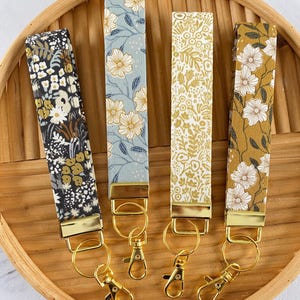 May include: Four floral key fobs in a wooden tray. The key fobs feature different floral patterns in black, blue, and gold. Each fob has a gold-colored metal clasp and ring for attaching keys.