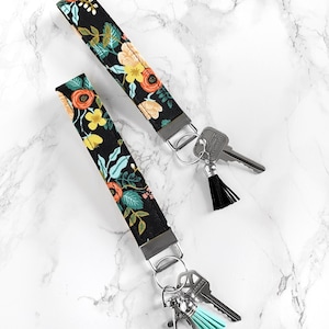 May include: Two black floral key wristlets with metal hardware and attached keys. The fabric features a colorful pattern of orange, yellow, and teal flowers and leaves. One wristlet has a black tassel, and the other has a teal tassel.