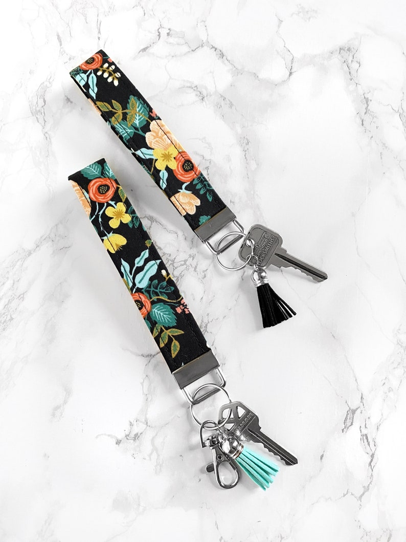 May include: Two black floral key wristlets with metal hardware and attached keys. The fabric features a colorful pattern of orange, yellow, and teal flowers and leaves. One wristlet has a black tassel, and the other has a teal tassel.