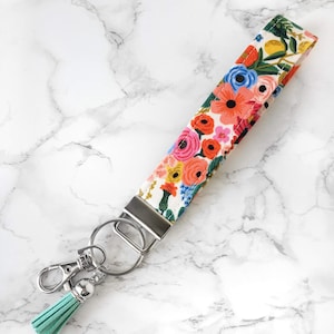 May include: A floral wristlet key fob with a colorful pattern of pink, orange, and blue flowers on a white background. It has a silver metal clasp and key ring, and a light teal tassel.
