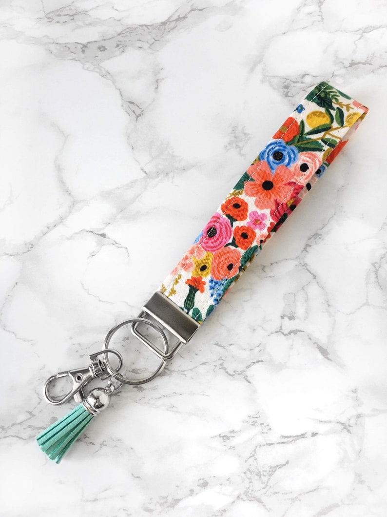 May include: A floral wristlet key fob with a colorful pattern of pink, orange, and blue flowers on a white background. It has a silver metal clasp and key ring, and a light teal tassel.