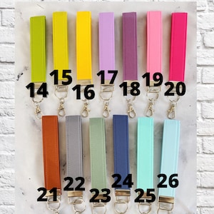 May include: A selection of fabric wrist keychains in various colors, including green, yellow, purple, pink, orange, gray, and blue. Each keychain has a metal clasp. The text "CHOOSE YOUR FABRIC" is at the top, and "SEE COLOR DESCRIPTIONS IN ITEM DETAILS" is at the bottom.