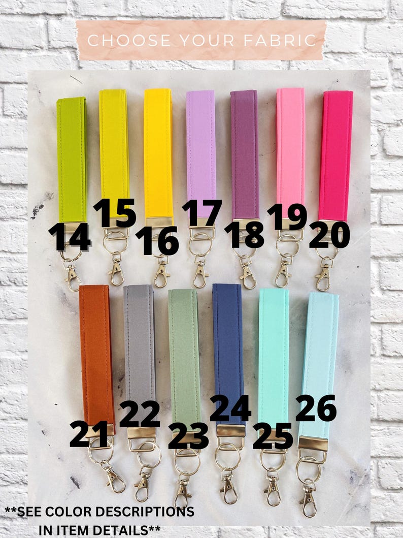 May include: A selection of fabric wrist keychains in various colors, including green, yellow, purple, pink, orange, gray, and blue. Each keychain has a metal clasp. The text "CHOOSE YOUR FABRIC" is at the top, and "SEE COLOR DESCRIPTIONS IN ITEM DETAILS" is at the bottom.