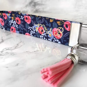 May include: A navy blue key fob with a floral pattern featuring pink and red roses, white flowers, and green leaves. The key fob has the embroidered text "Mrs. B" in gold thread and a pink tassel.