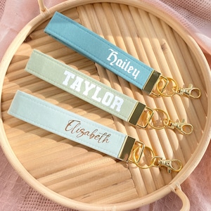 May include: Three personalized wrist keychains in pastel colors: light blue, sage green, and mint. Each keychain features a name in a contrasting font: "Hailey", "Taylor", and "Elizabeth". Gold-colored hardware and a swivel clasp complete the design. The keychains are displayed on a woven tray.