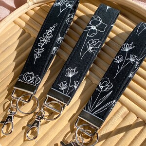May include: Three black wrist keychains with white floral designs. Each keychain has a silver-colored metal clasp and ring. The keychains are arranged on a light brown woven tray, with a pink fabric in the background.