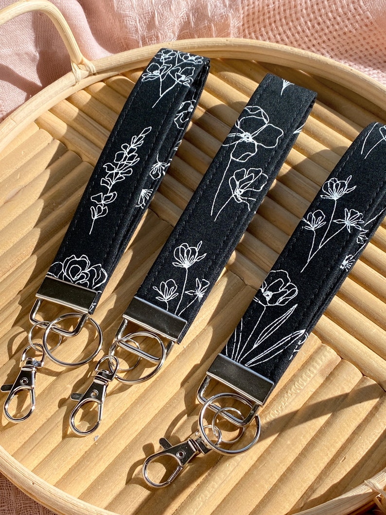 May include: Three black wrist keychains with white floral designs. Each keychain has a silver-colored metal clasp and ring. The keychains are arranged on a light brown woven tray, with a pink fabric in the background.