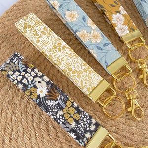May include: Close-up of several floral patterned key fobs with gold-colored hardware. The key fobs feature various designs, including white and yellow flowers on a blue background, and a black and white floral pattern. They are arranged on a woven surface.