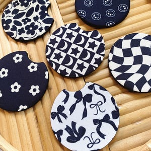 May include: A collection of six circular car coasters with various black and white patterns. Designs include flowers, smiley faces, stars and moons, bows, and a checkered pattern. The coasters are arranged on a woven, light brown tray.