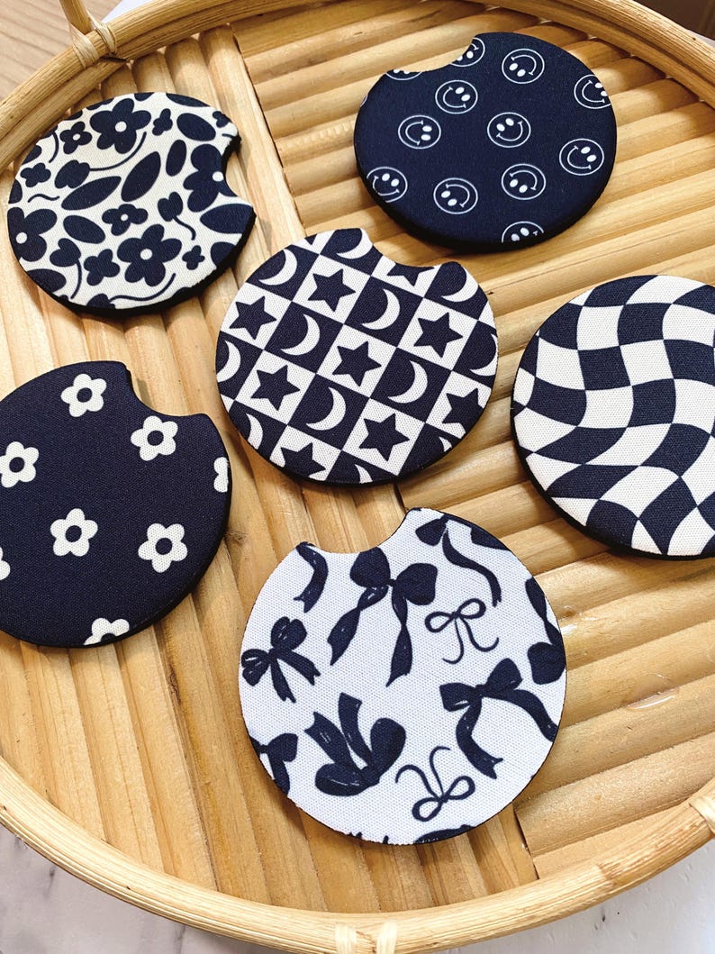 May include: A collection of six circular car coasters with various black and white patterns. Designs include flowers, smiley faces, stars and moons, bows, and a checkered pattern. The coasters are arranged on a woven, light brown tray.